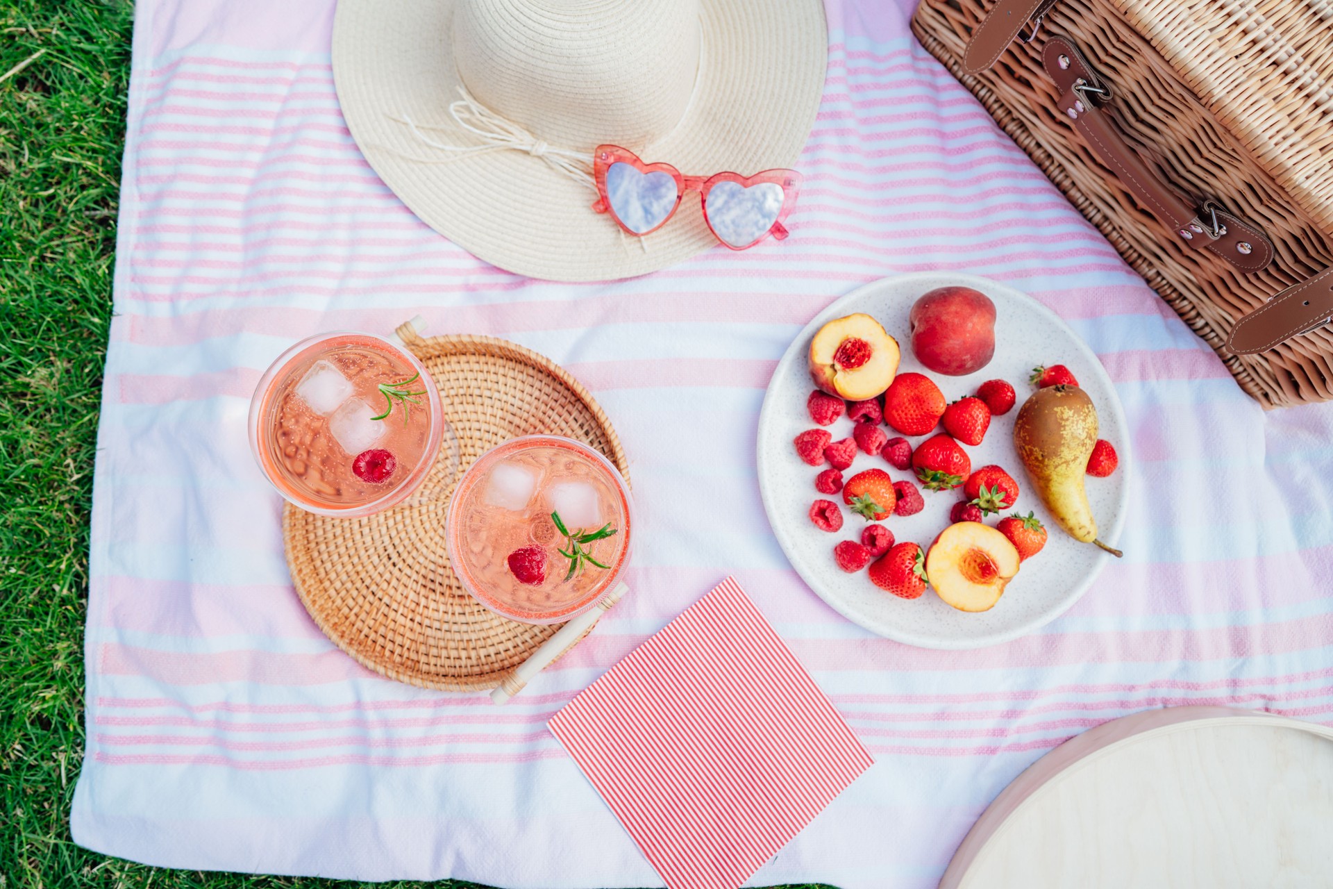 Top view pink drinks, cocktail with ice, raspberry, rosemary. Two glasses with martini, champagne, cider, lemonade on the tray on blanket with fruit plate, picnic basket. Cozy summer picnic on nature